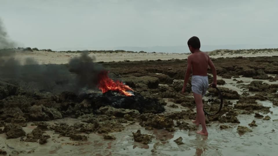 A boy walks across a rocky shoreline past a burning tyre