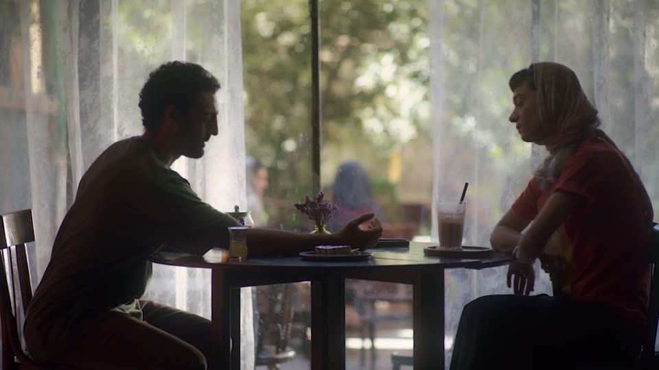A man and a woman talk across a café table by a lace-curtained window
