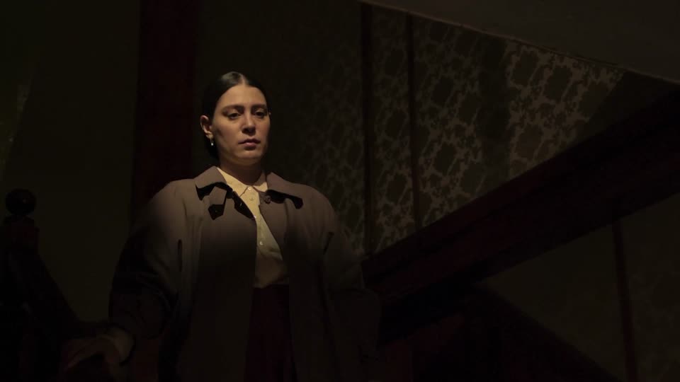 A woman in a trench coat stands at the foot of a dark stairwell